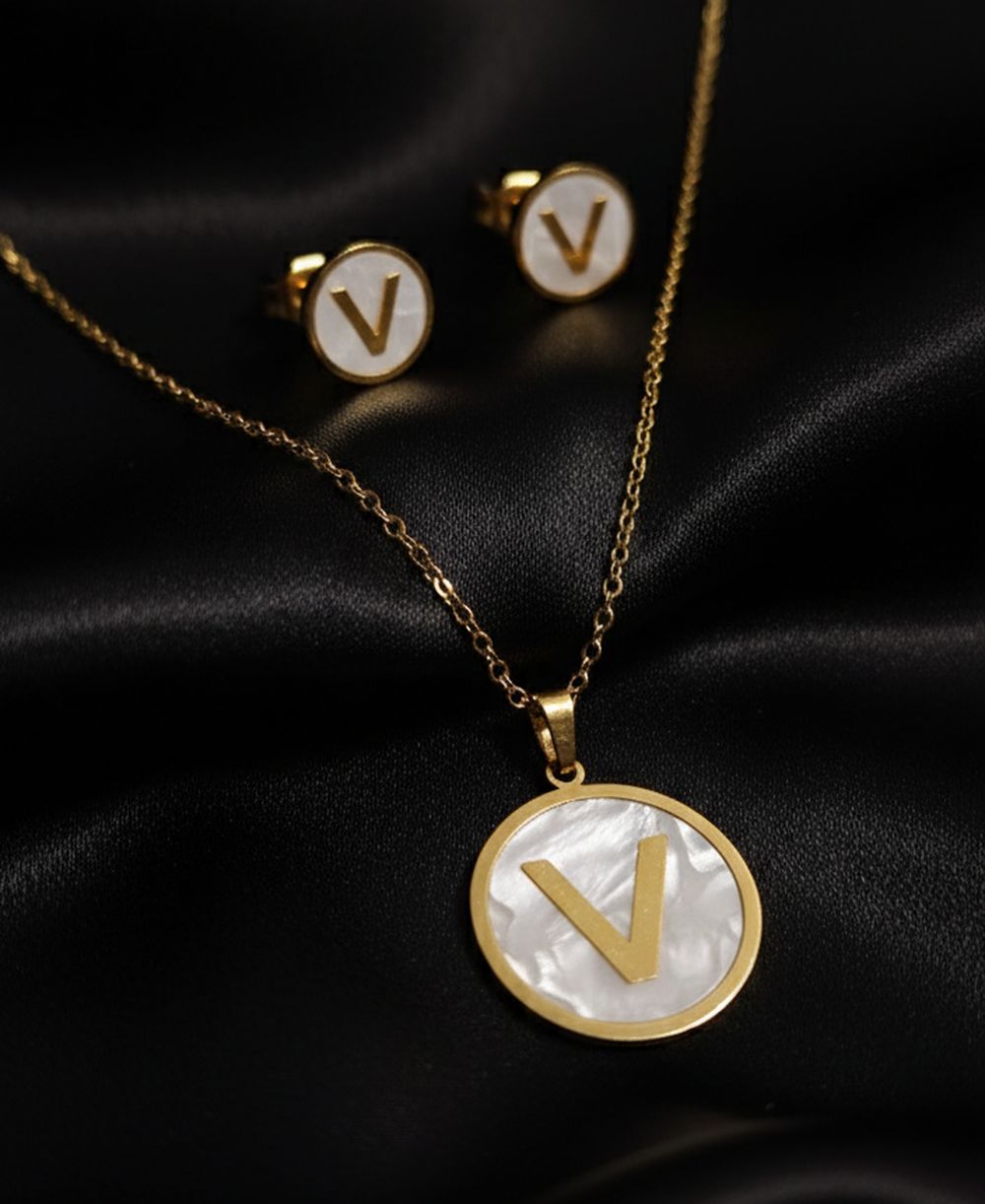 Golden initial “V” necklace and matching white enamel stud earrings on a dark background