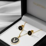 Golden initial “V” necklace and matching black enamel stud earrings on a dark background