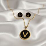 Golden initial “V” necklace and matching black enamel stud earrings on a dark background