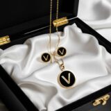 Golden initial “V” necklace and matching black enamel stud earrings on a dark background
